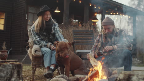 Young-Couple-and-Dog-Sitting-by-Bonfire-near-Farmhouse